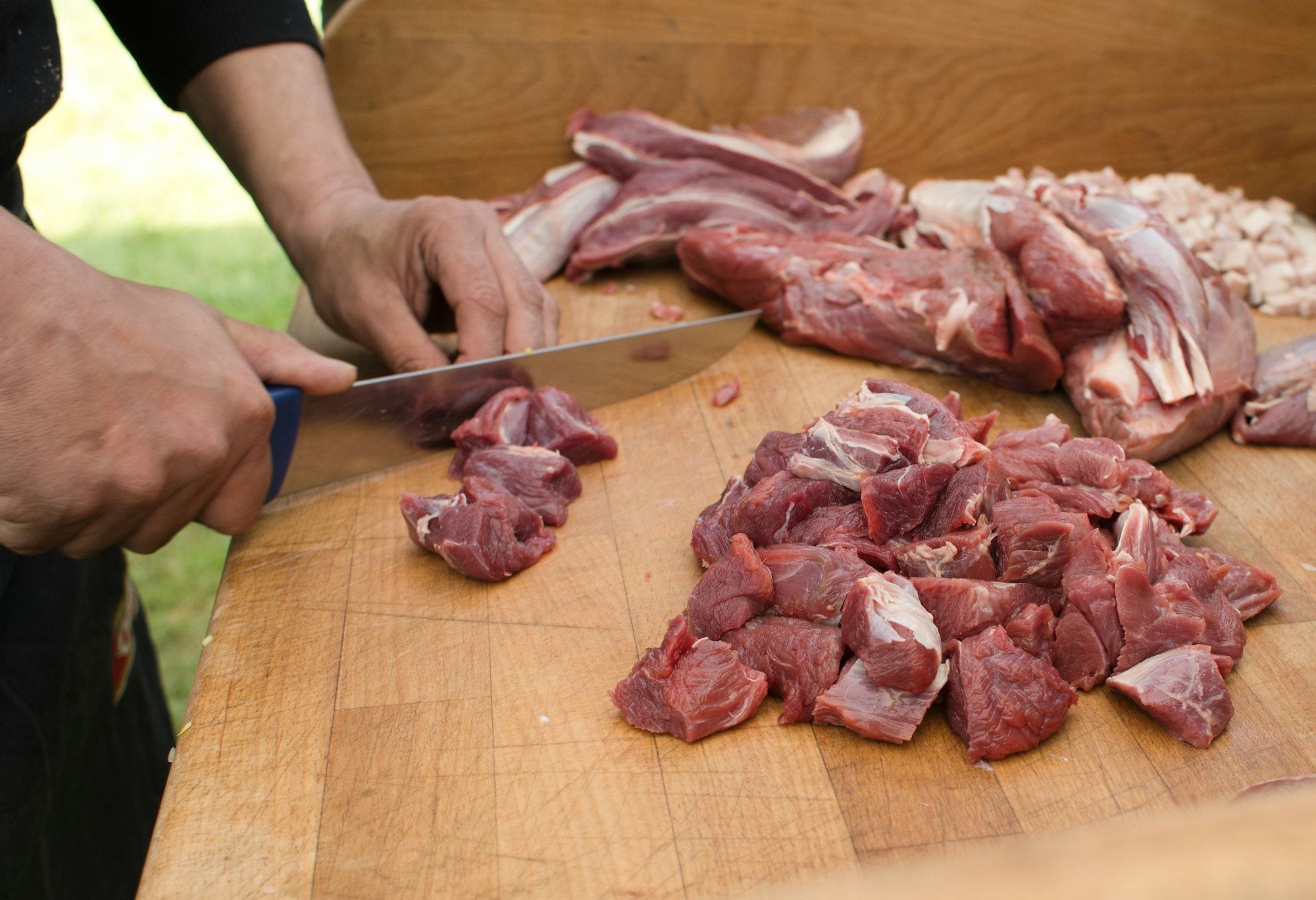 A butcher's hands cutting fresh red meat into curry-cut pieces on a wooden block